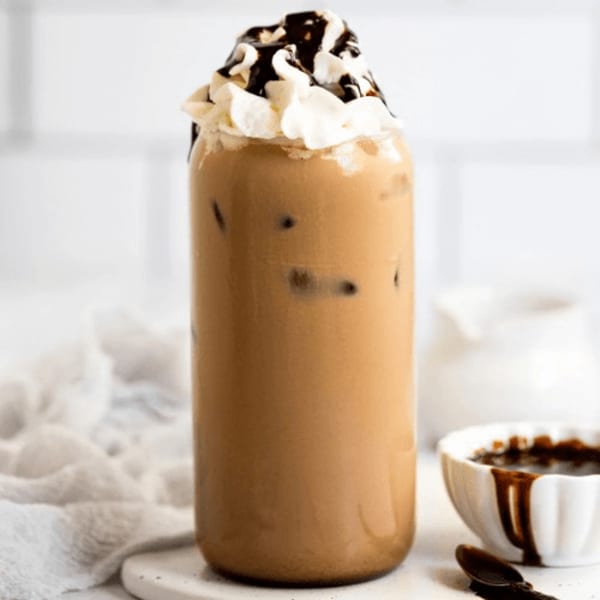 Iced coffee with whipped cream and chocolate drizzle, next to a bowl of chocolate sauce.