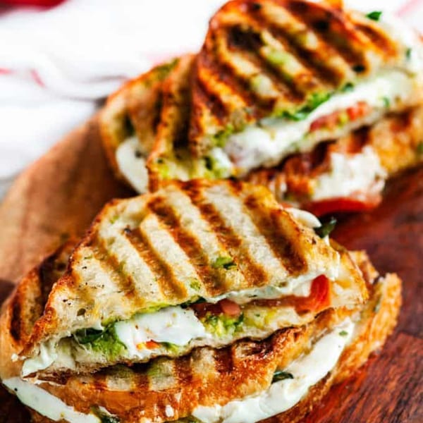 Two grilled paninis with melted cheese, tomatoes, and green filling on a wooden board.