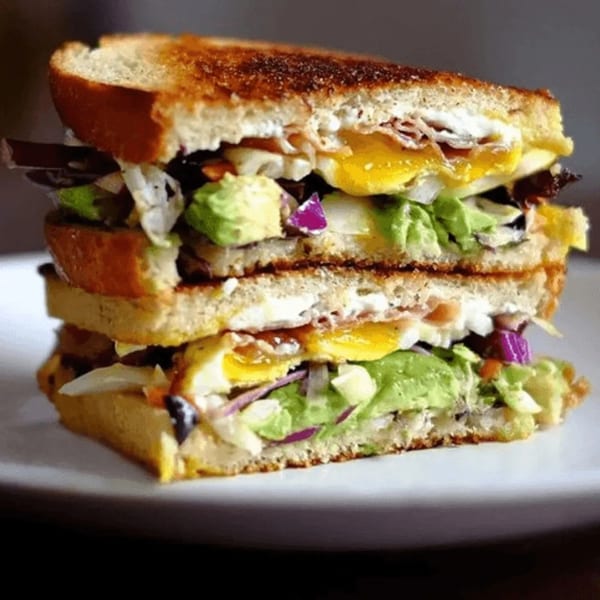 A stacked breakfast sandwich with fried eggs, avocado, and red onion on toasted bread.
